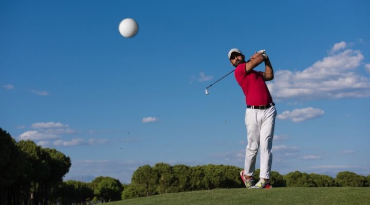 A golfer in red has just swung his club, sending the golf ball spinning mid-air